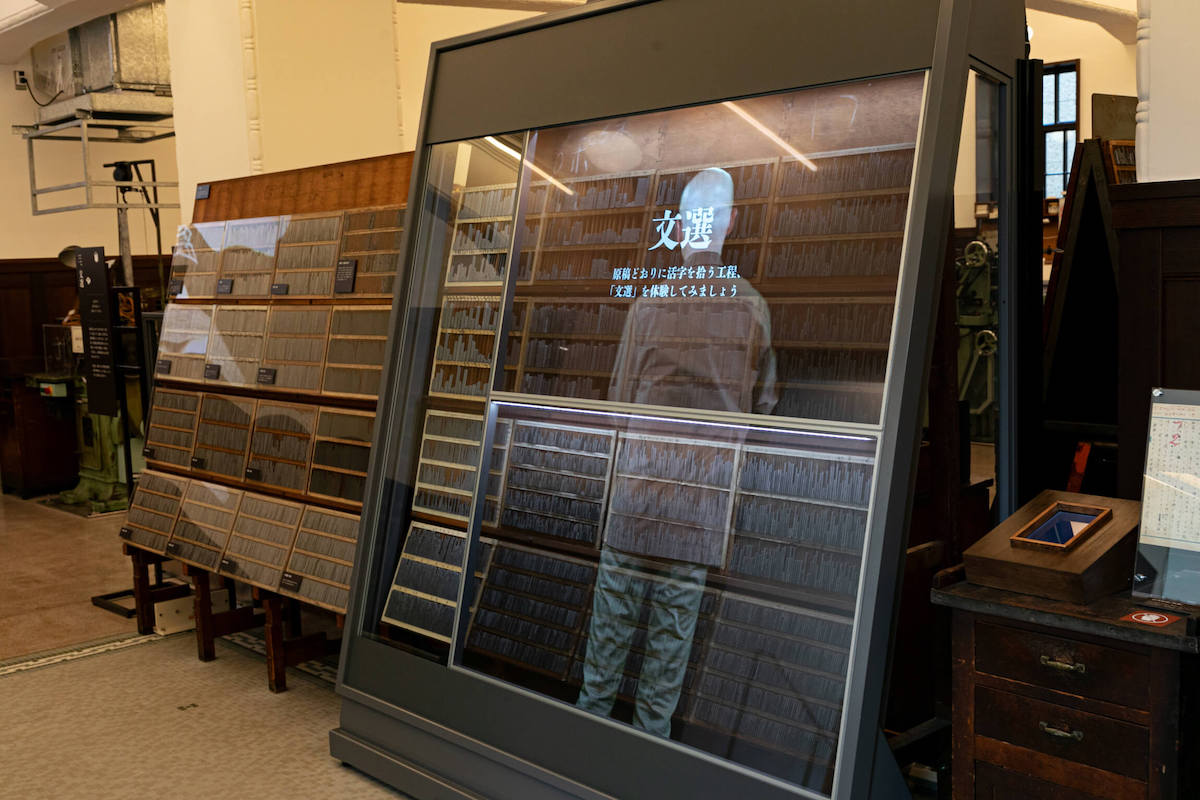 透明モニターには職人が文選するシーンが映し出される。文字の大きさや書体が指定された原稿と文選箱という活字を入れる箱を持ち、拾っていく