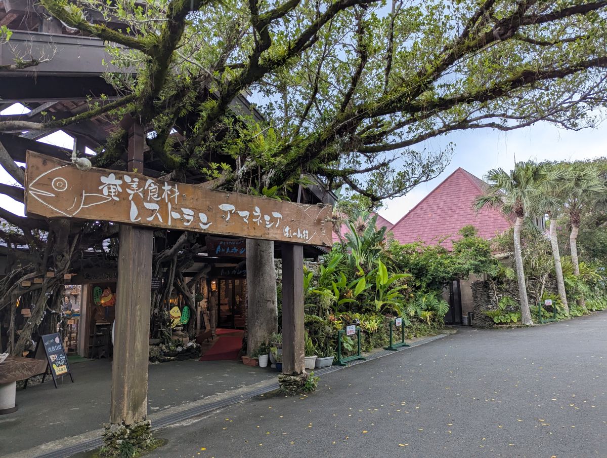 入り口に立つ大きなシンボルツリー。 木の看板には大きな魚が描かれている。 右側にレストランAMAネシア、中央にショップがある。